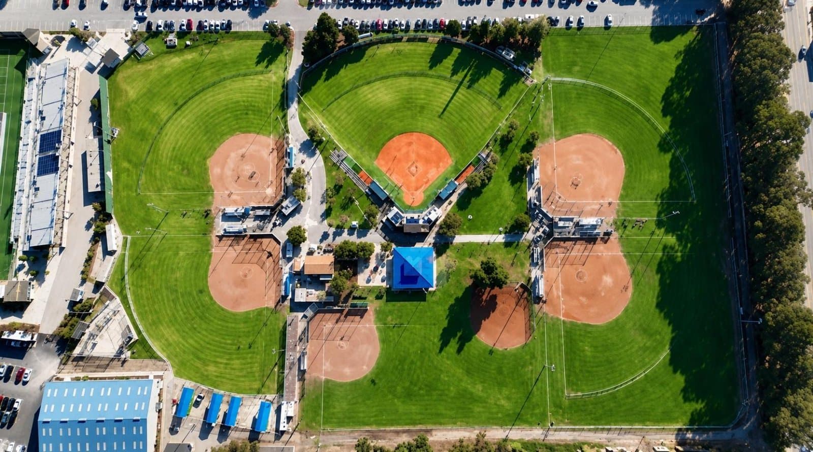 Overhead view of the Salinas Sports Complex softball facilities, showing four diamonds arranged in a cloverleaf around a central hub building, with outfields of mown grass and parking beyond.